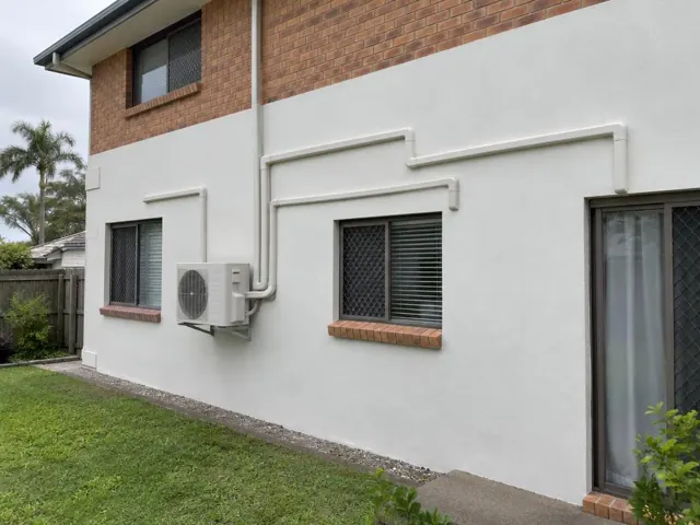 Outdoor split-system unit with pipe covers on a two-storey brick and rendered house wall in Carrara